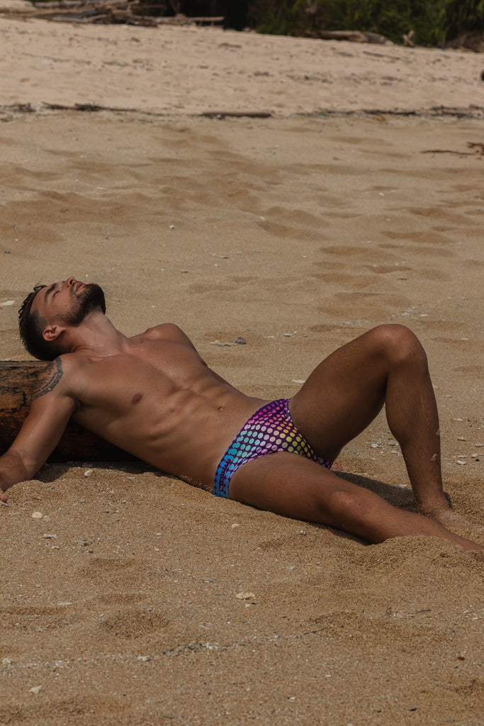A sandy beach in Australia is the setting for a man who lies backward with his head resting on a log. His eyes are closed and he faces up towards the sky. One knee is bent and his arms are out beside him. He is very fit with his abs the focus and all he wears is rainbow pride swim trunks that mimic the classic speedo. The swimsuit is purple with rainbow polka dots.